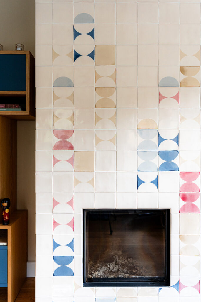 Fireplace surround of cream Smink Studio handmade ceramic tiles with blue, pink and tan half-circle screen-printed colours forming vertical accents around a black firebox.