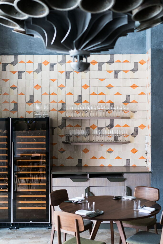 Smink Studio handmade ceramic tiles in cream, orange and grey geometric screen-printed colours lining a restaurant wall with wine storage, glassware shelves and dining tables.