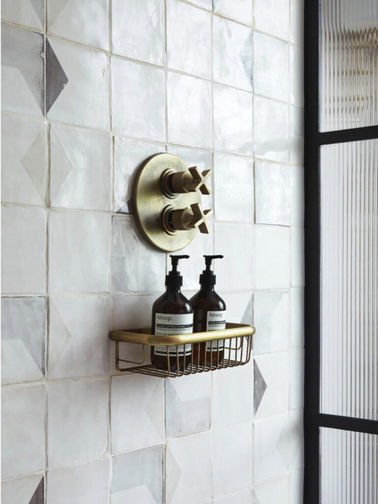 Shower wall of white Smink Studio handmade ceramic tiles with soft grey diamond screen-printed colours, brass shower valve and basket, and a fluted glass screen.