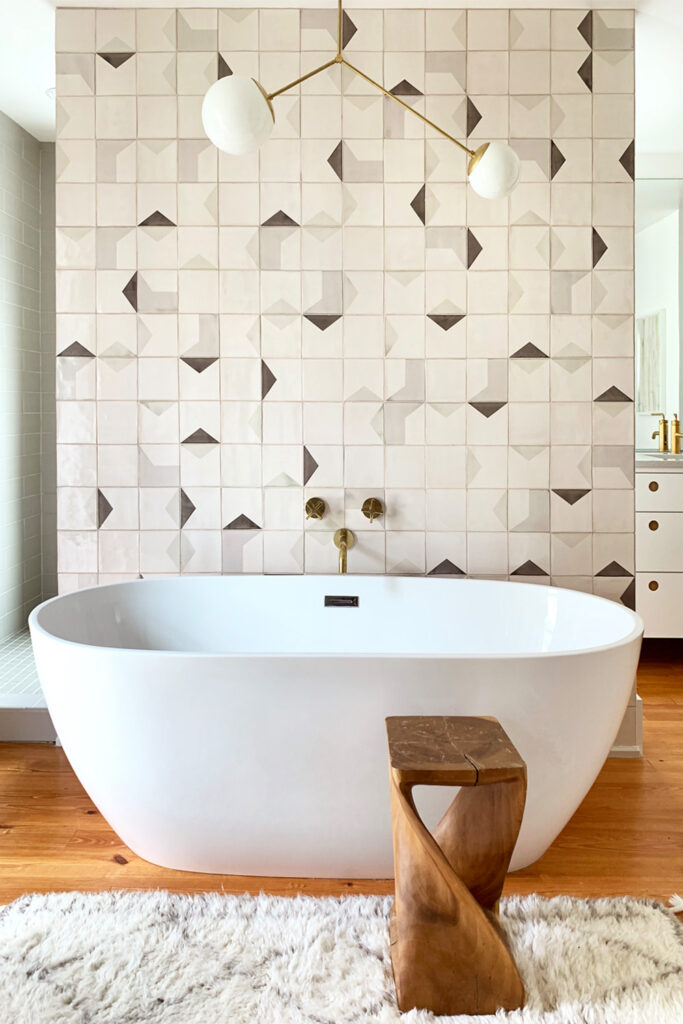 Freestanding white bath against a feature wall of Smink Studio handmade ceramic tiles in cream and taupe geometric screen-printed colours, with brass fittings and a pendant light.