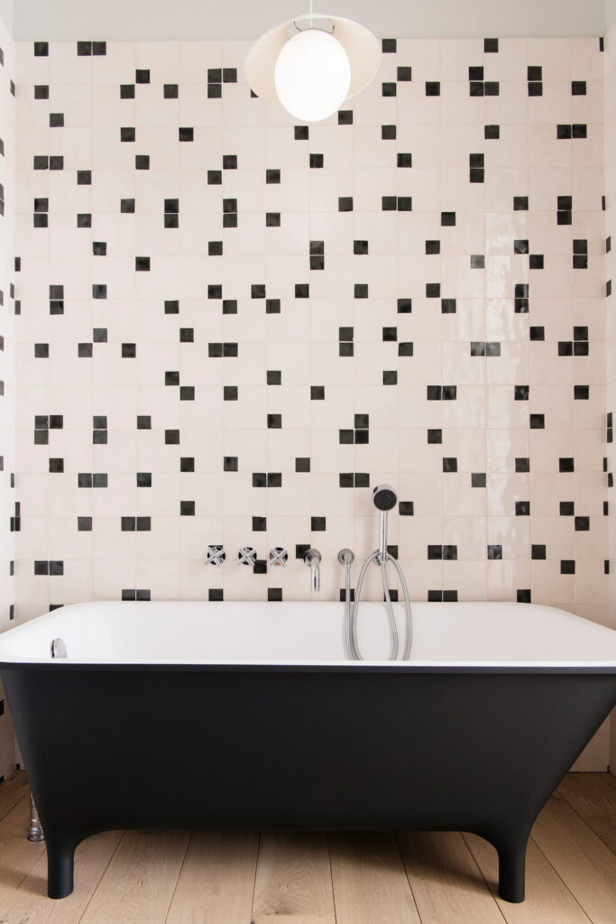 Black roll-top bath against blush-pink Smink Studio handmade ceramic tiles scattered with black squares in screen-printed colours.