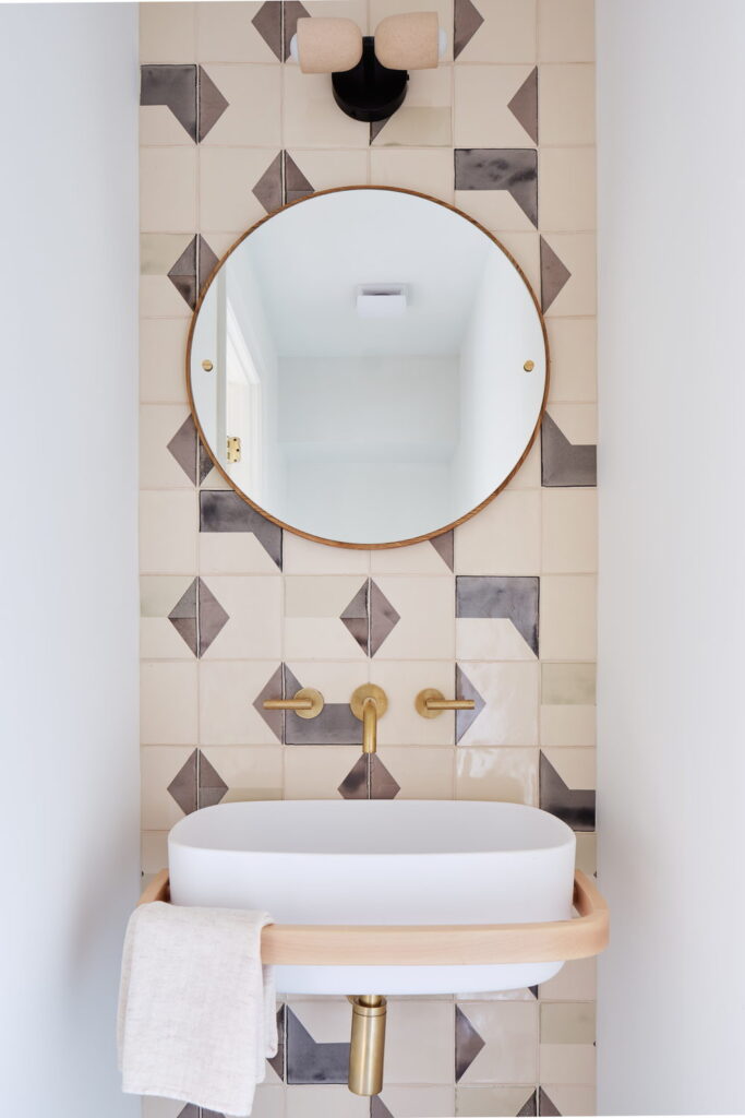 Powder room feature wall of Smink Studio handmade ceramic tiles in cream and charcoal geometric screen-printed colours, with a round mirror, white basin and brass taps.