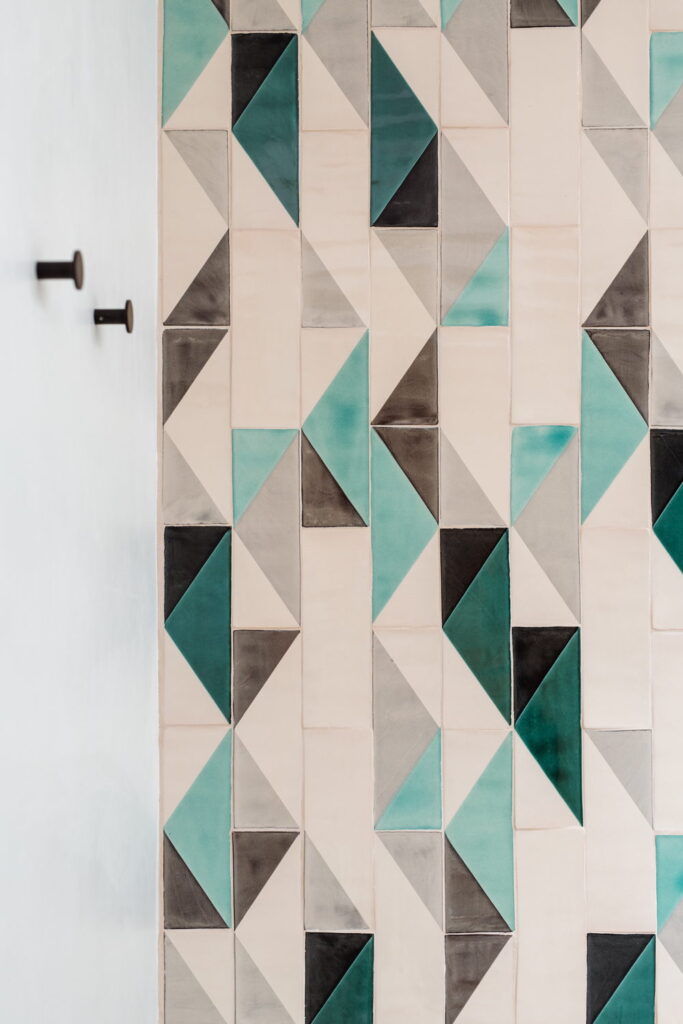 Detail of Smink Studio handmade ceramic tiles in cream, grey and turquoise triangle pattern with screen-printed colours, fitted with two black wall hooks.