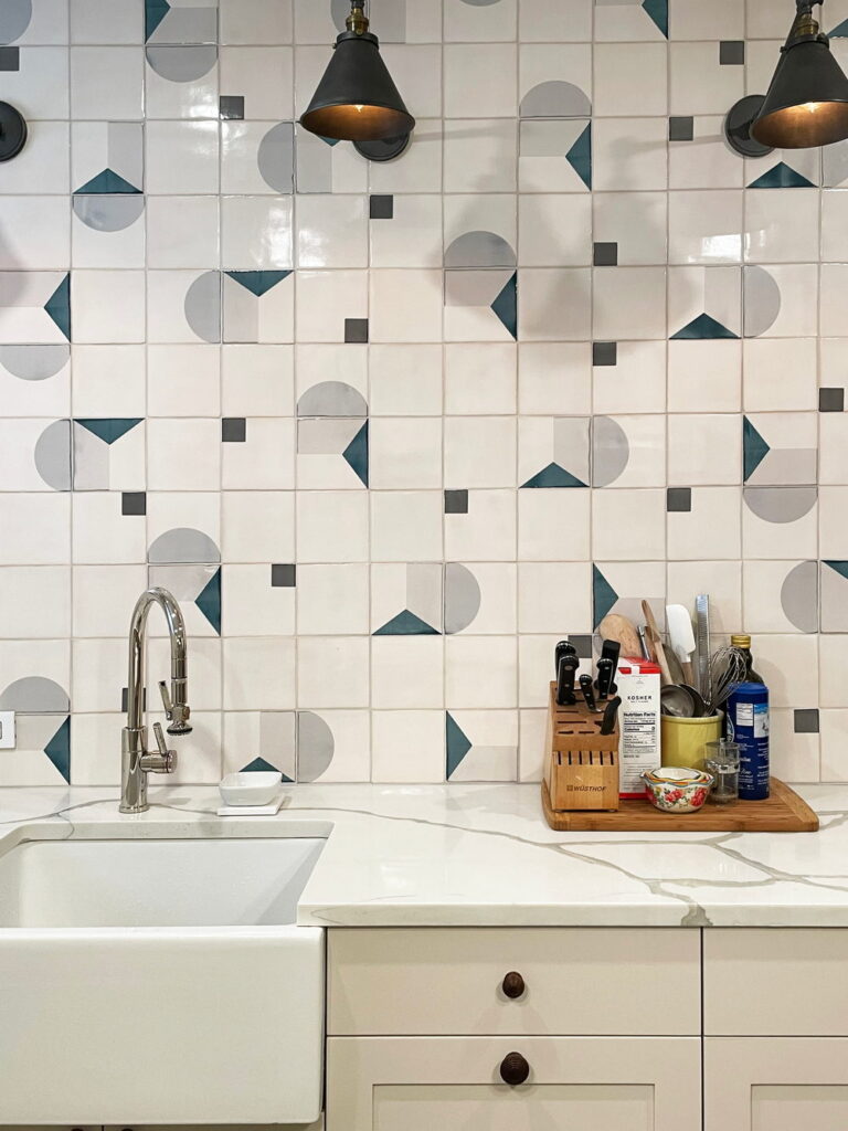 Kitchen backed by white Smink Studio handmade ceramic tiles with grey and teal circle and triangle screen-printed colours, marble counter and farmhouse sink.