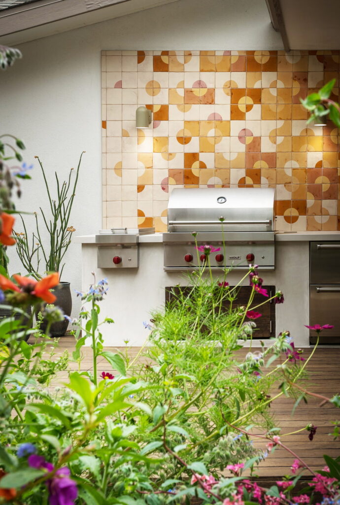 Outdoor kitchen backed by Smink Studio handmade ceramic tiles in cream, orange, terracotta and pink half-circle screen-printed colours, behind a grill with garden flowers in front.