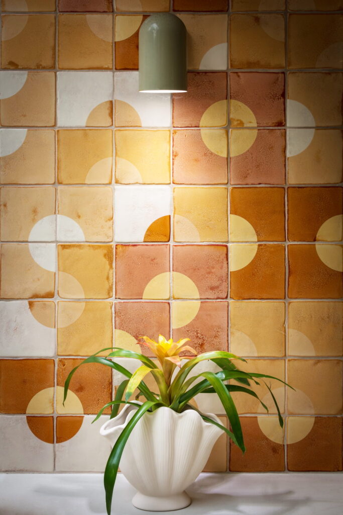 Detail of warm amber, terracotta and cream Smink Studio handmade ceramic tiles with half-circle screen-printed colours, lit by a sage pendant, with a white vase.