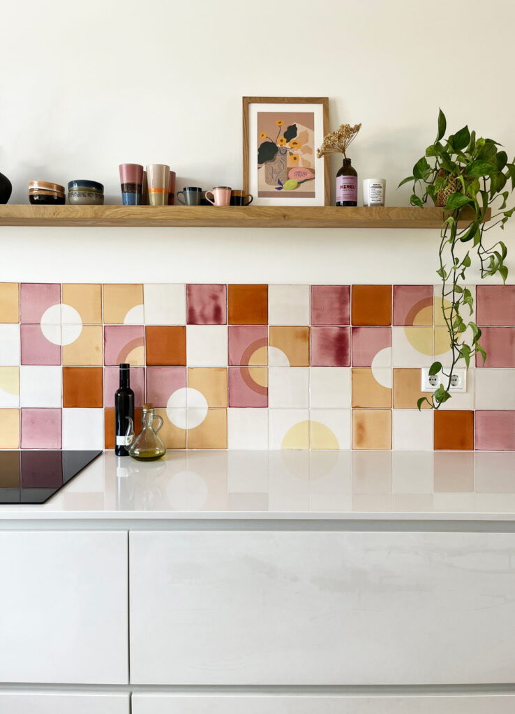 Kitchen splashback of Smink Studio handmade ceramic tiles in pink, terracotta, mustard and cream half-circle screen-printed colours, with a white counter and oak shelf.
