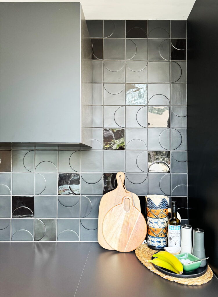 Kitchen backed by grey and charcoal Smink Studio handmade ceramic tiles with half-circle screen-printed colours and metallic-lustre accents, beside a dark counter.