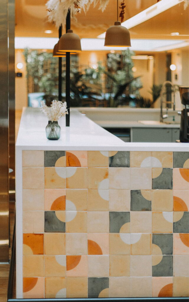 Cafe counter clad in Smink Studio handmade ceramic tiles in cream, apricot, grey and orange half-circle screen-printed colours, with dome pendants overhead.