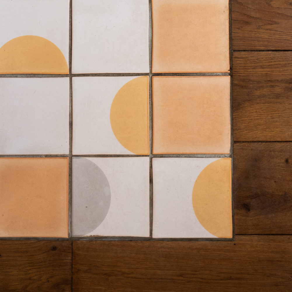 Detail of Smink Studio handmade ceramic tiles in cream, apricot, grey and orange half-circle screen-printed colours, beside oak floorboards.
