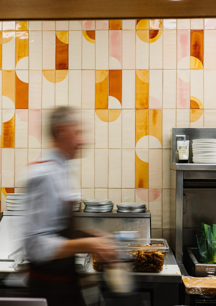 Restaurant kitchen backed by Smink Studio handmade ceramic tiles in cream, orange and pink half-circle screen-printed colours, with a chef in motion at the pass.