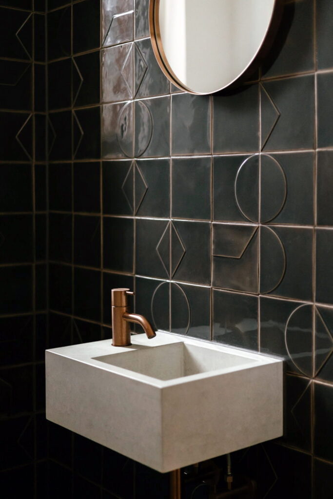 Powder room of charcoal Smink Studio handmade ceramic tiles with embossed geometric line detailing, with a concrete basin, copper tap and round mirror.