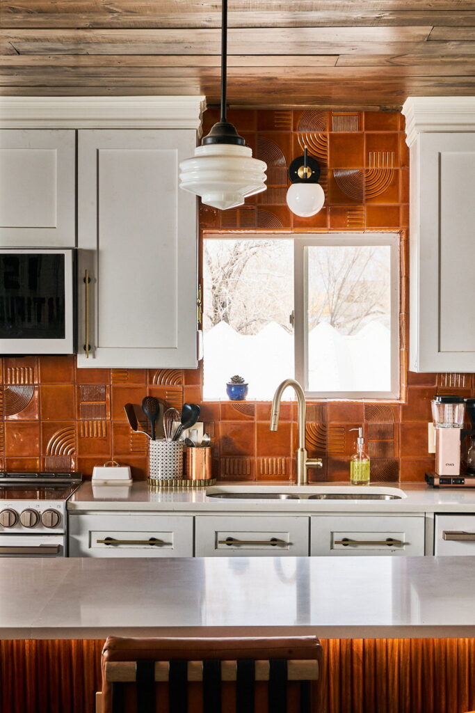 Kitchen of terracotta Smink Studio handmade ceramic tiles with embossed arch and line detailing around a window, with white cabinets and a brass tap.