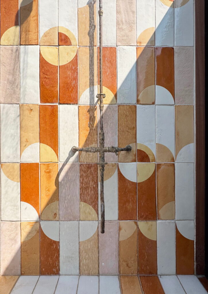 Outdoor shower of Smink Studio handmade ceramic tiles in terracotta, orange, mustard and cream half-circle screen-printed colours, with an aged brass shower in sunlight.