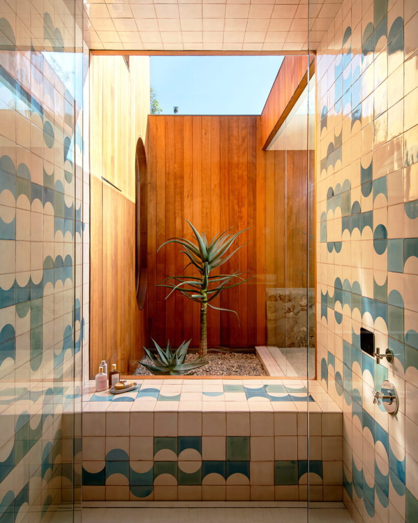 Courtyard shower of blue-green and cream Smink Studio handmade ceramic tiles with half-circle screen-printed colours, looking through glass to a timber-clad courtyard.