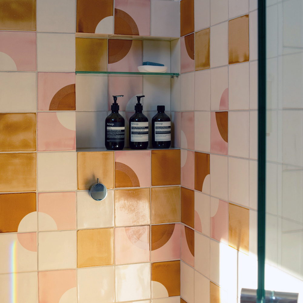 Shower corner of Smink Studio handmade ceramic tiles in cream, pink, orange and mustard half-circle screen-printed colours, with a niche of bottles behind a glass screen.