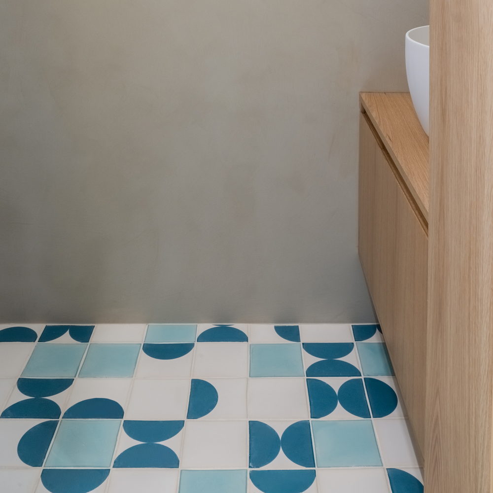 Powder room floor of white, teal and blue Smink Studio handmade ceramic tiles with half-circle screen-printed colours, beside an oak vanity and white basin.