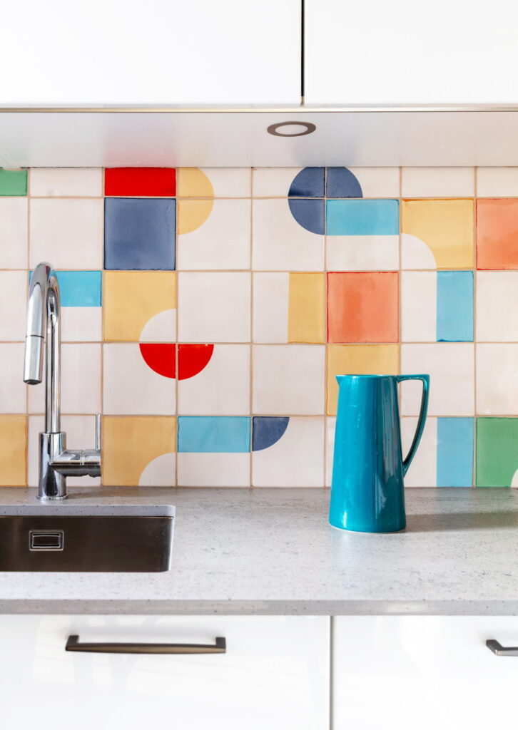 Kitchen splashback of white Smink Studio handmade ceramic tiles with bold red, blue, orange, teal and green square and half-circle screen-printed colours, with a teal jug.
