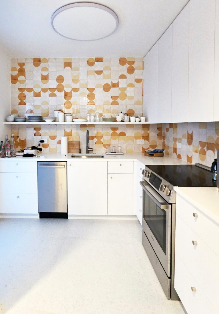 Kitchen backed by Smink Studio handmade ceramic tiles in cream, mustard and tan half-circle screen-printed colours, with white cabinets and a stainless range.